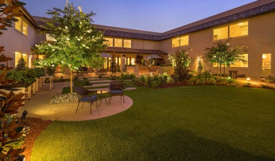 Well-lit courtyard at dusk with chairs, tables, planted trees, and a surrounding two-story building.