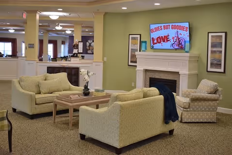 A cozy living room area in a senior living facility with two light green sofas, a patterned armchair, a wooden coffee table with decorative items, and a fireplace with a TV mounted above it displaying the message 'OLDIES BUT GOODIES LOVE'. The walls are painted light green and decorated with framed artwork.