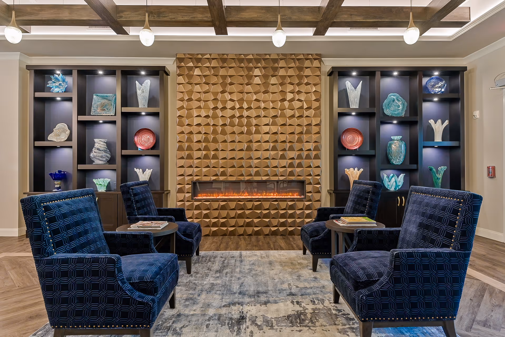 A cozy sitting area with four dark blue upholstered armchairs arranged around two small round tables. Behind the chairs is a modern electric fireplace set in a textured geometric wooden wall. On either side of the fireplace are built-in shelves displaying various decorative glass art pieces. The ceiling features wooden beams with hanging pendant lights, and the floor has a patterned rug over wood flooring.