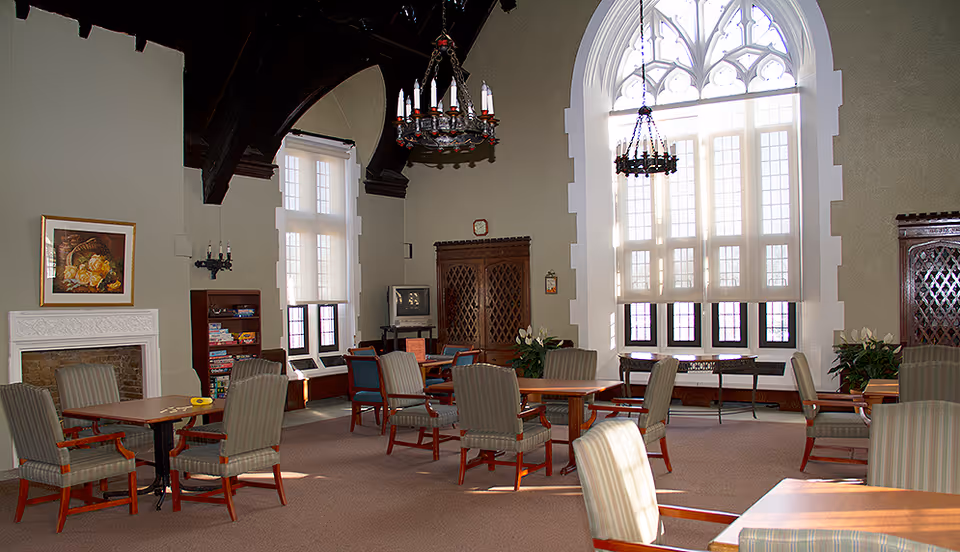 Spacious bright common room with high arched windows, chandeliers, and multiple tables and upholstered chairs.