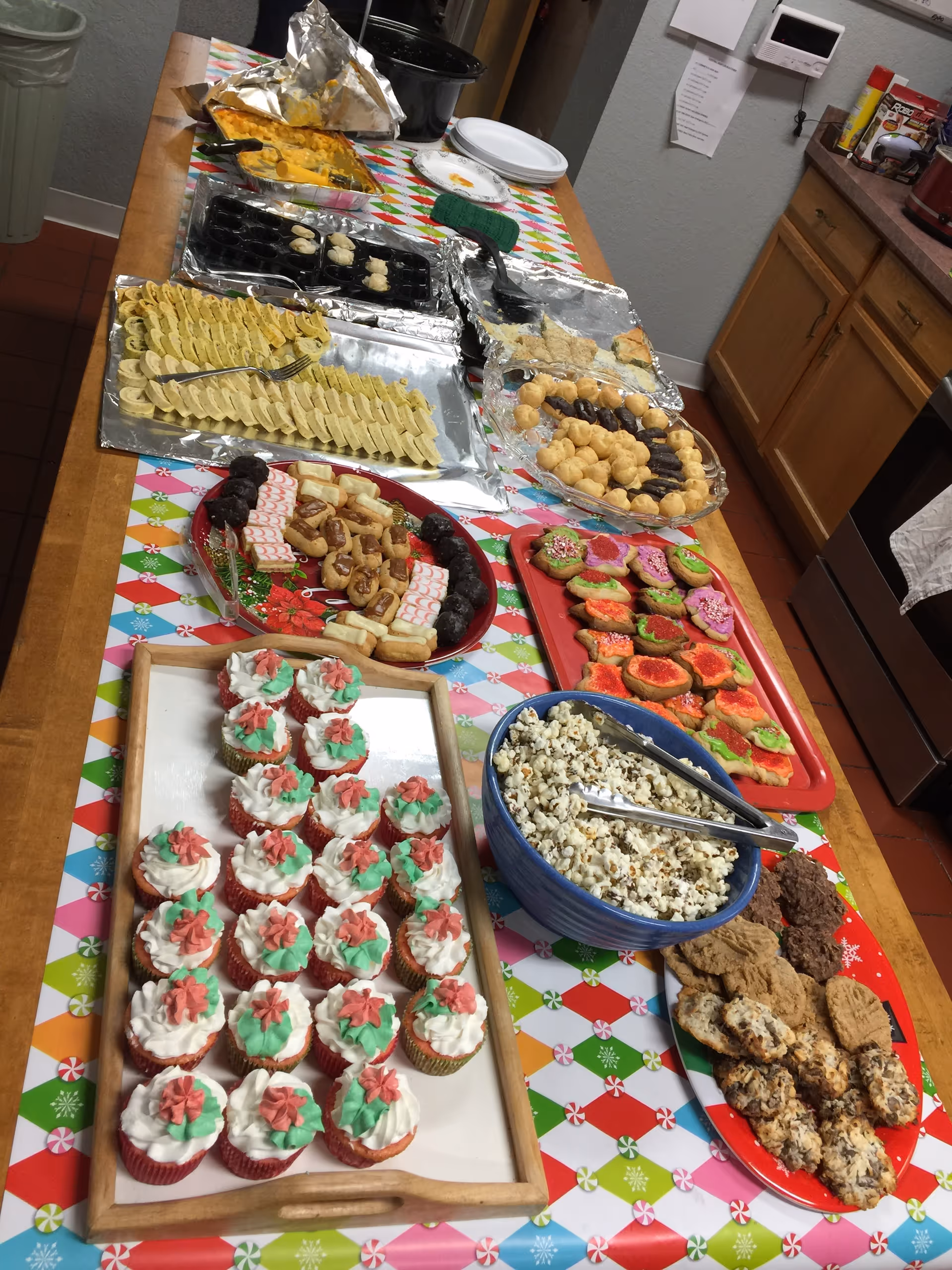 Long kitchen table covered with a festive tablecloth holding a buffet of desserts and snacks including cupcakes, cookies, popcorn and pastries.