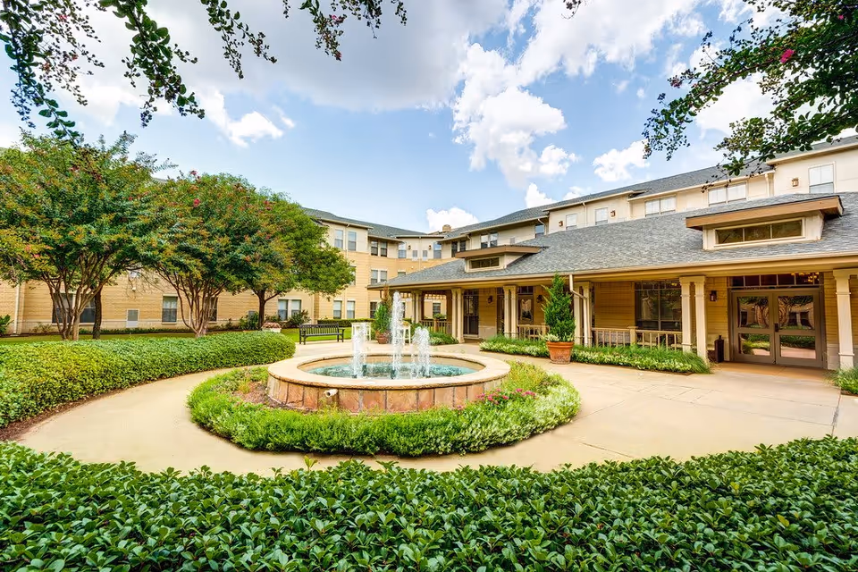 Outdoor courtyard area of Town Village Crossing senior living facility featuring a circular fountain with water jets in the center, surrounded by green shrubs and a paved walkway. The building has a beige exterior with multiple windows and a covered entrance. Trees and benches are visible along the walkway under a partly cloudy sky.