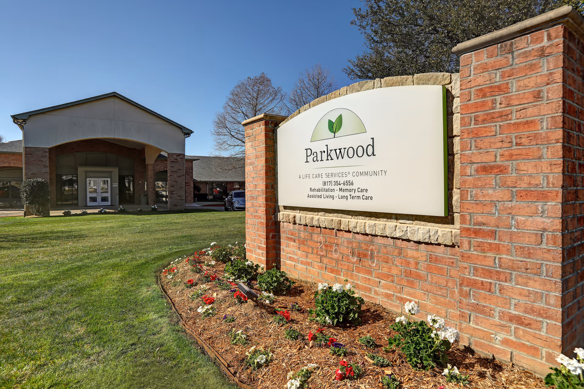 Exterior view of the Parkwood Healthcare facility showing a brick sign with the facility name and services offered, a well-maintained lawn, flower bed, and the building entrance under a clear blue sky.