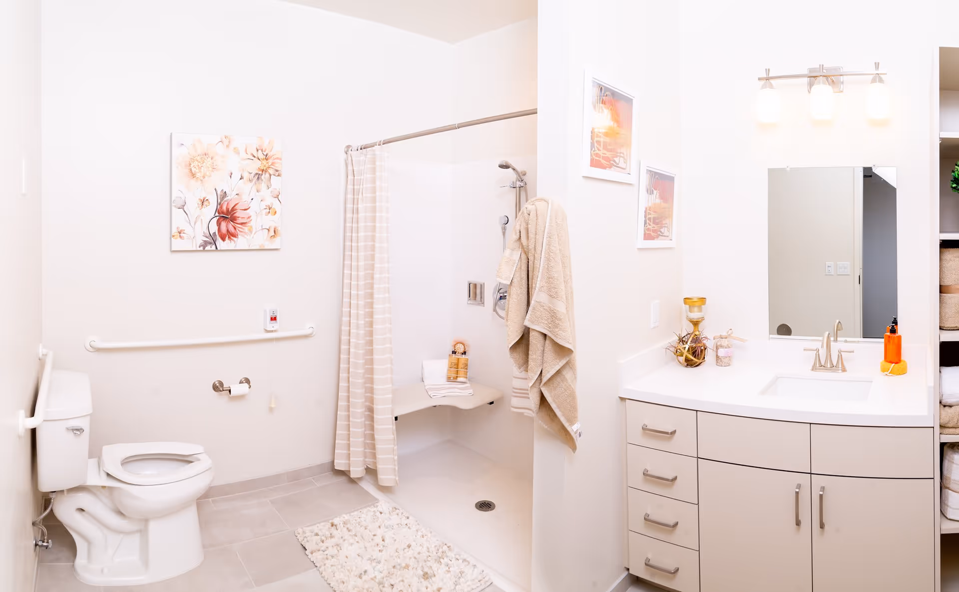 A bright and clean bathroom featuring a toilet with grab bars, a walk-in shower with a striped curtain and a built-in seat, beige towels hanging on the wall, a white vanity with a sink, mirror, and light fixture above, and decorative artwork on the walls.