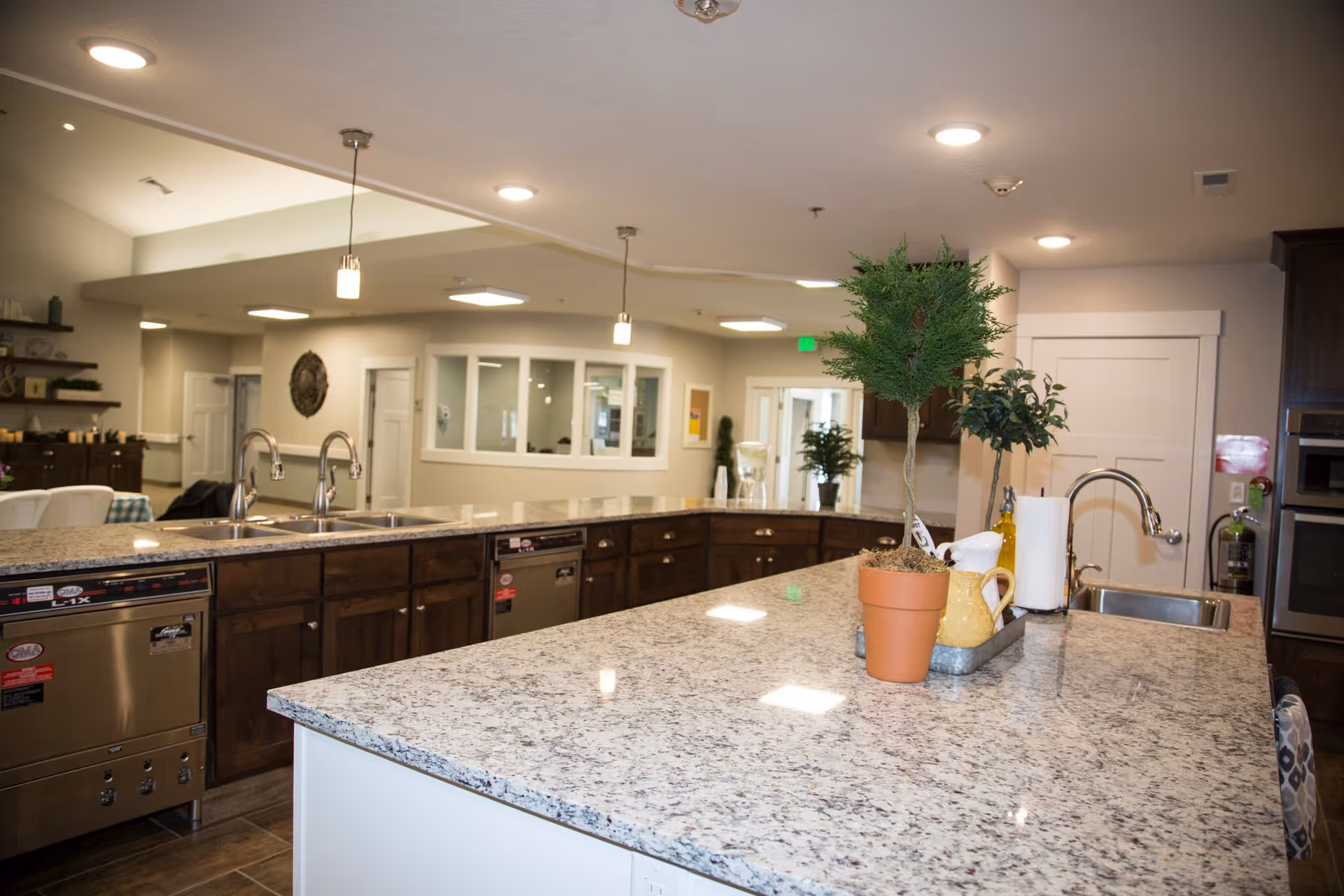 A spacious kitchen area in a senior living facility with granite countertops, two sinks with chrome faucets, and dark wooden cabinets. There are potted plants and kitchen items on the counter, with bright ceiling lights illuminating the space. In the background, there are windows, doors, and a dining area with chairs and tables.