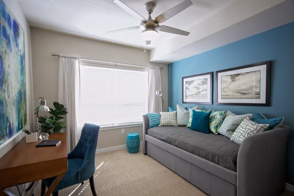 A cozy living room with a gray daybed covered with multiple patterned pillows in shades of blue, green, and white. The wall behind the daybed is painted blue and has two framed black and white photographs. A large window with white curtains lets in natural light. A wooden desk with a blue upholstered chair is positioned against the opposite wall, accompanied by a desk lamp and a small plant. The room has a ceiling fan with a light fixture.