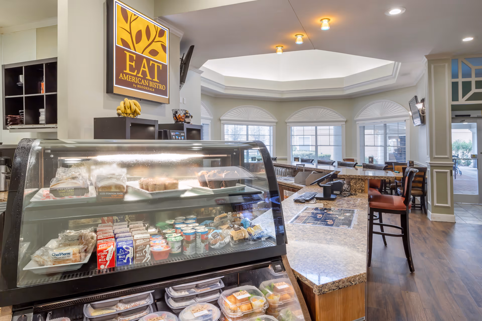 Interior view of a dining area at Brookdale Clear Lake featuring a glass display case with various packaged food items and beverages. Behind the display case is a counter with bar stools and a sign that reads 'EAT American Bistro by Brookdale'. The room has large windows with blinds and a high ceiling with recessed lighting.