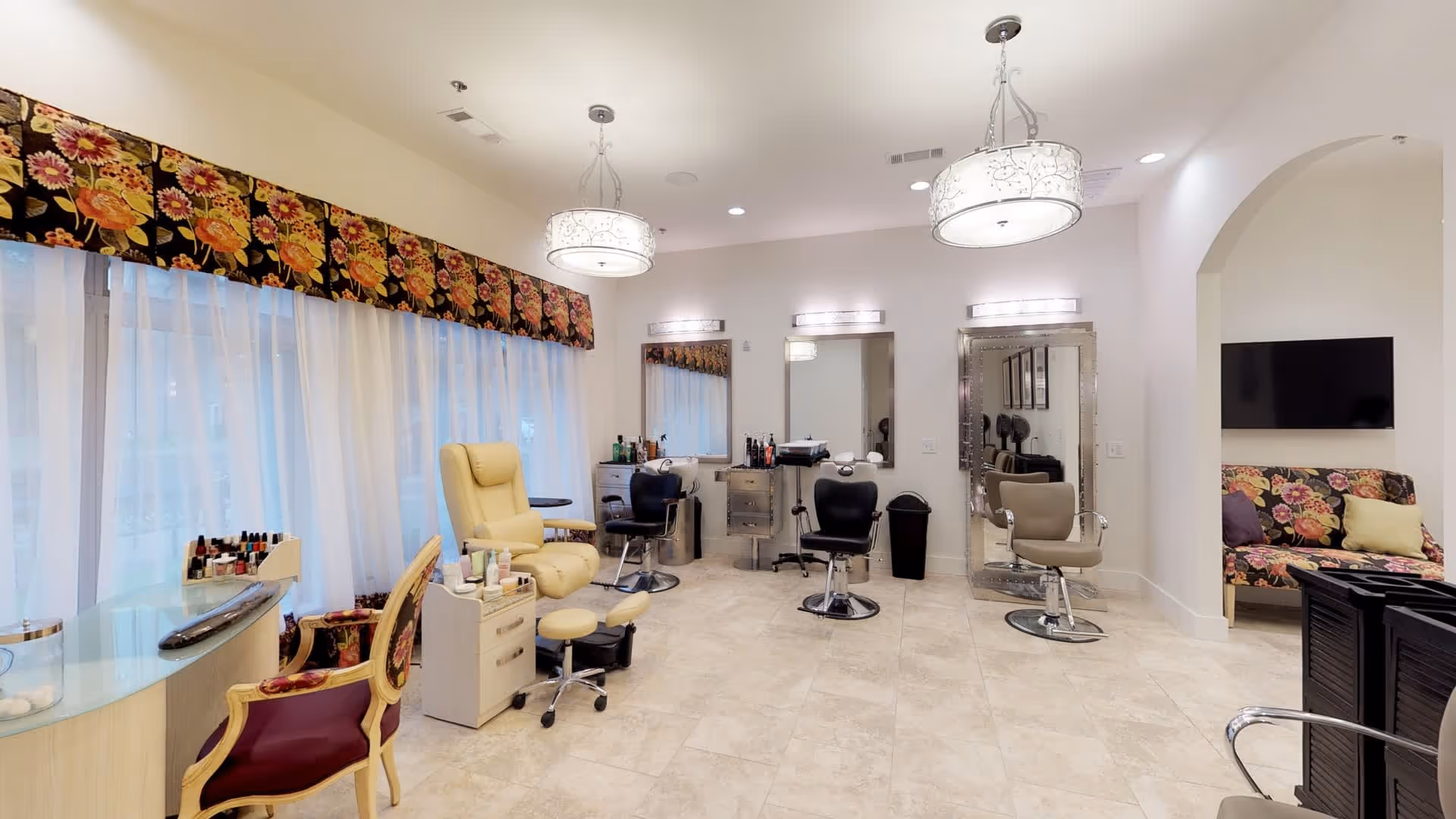 A bright and spacious salon area with multiple styling chairs and mirrors. There is a comfortable yellow pedicure chair with footrests, a manicure station with nail polish bottles, and several black salon chairs in front of large mirrors. The room has large windows with sheer white curtains and floral valances. In the background, there is a floral-patterned couch and a wall-mounted TV.