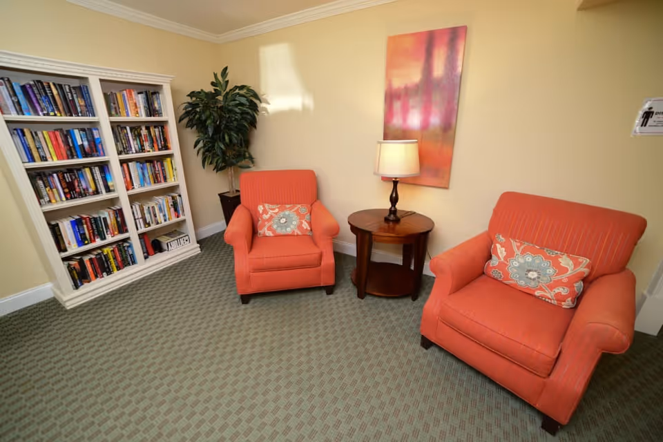 A cozy sitting area in a senior living facility with two orange armchairs featuring patterned cushions, a round wooden side table with a lamp, a tall white bookshelf filled with books, a potted plant, and a colorful abstract painting on the wall.