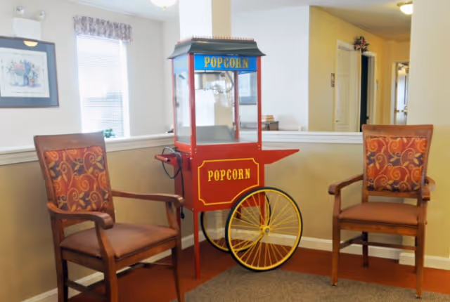 A popcorn machine on wheels positioned between two wooden chairs with patterned cushions in a well-lit room. The room has beige walls, a window with a floral valance, a framed picture on the wall, and a hallway visible in the background.