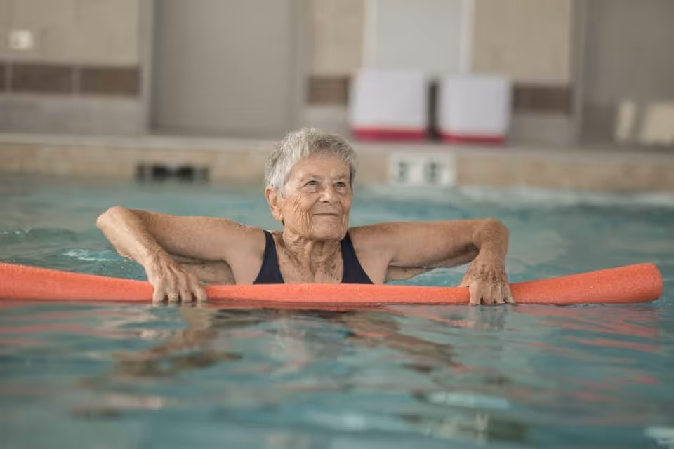 An elderly woman in a black swimsuit holding a red pool noodle while standing in an indoor swimming pool.