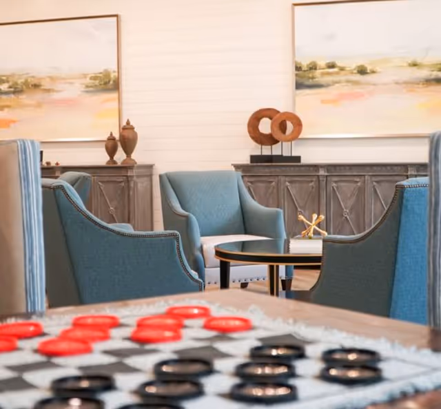 Seating area with teal armchairs around a round coffee table, artwork and cabinets in the background, and a checkers board in the foreground.