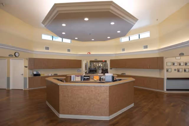 Interior view of a reception or nurse station area in a senior living facility with a large octagonal desk in the center, surrounded by wooden cabinets and a hallway in the background. The ceiling features recessed lighting and a raised octagonal design with windows near the top of the walls allowing natural light.