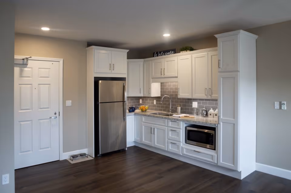 Modern kitchen area with white cabinets, a stainless steel refrigerator, a built-in microwave, a sink, and a gray tiled backsplash. The floor is dark wood, and there is a white door to the left. The kitchen is well-lit with recessed ceiling lights.