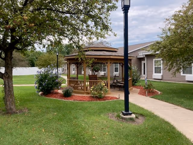 Outdoor area of Windsor Manor featuring a wooden gazebo surrounded by green grass, trees, and landscaped flower beds with mulch. A paved walkway leads to the gazebo and a nearby building with beige siding and white-trimmed windows. A black lamp post stands near the walkway.