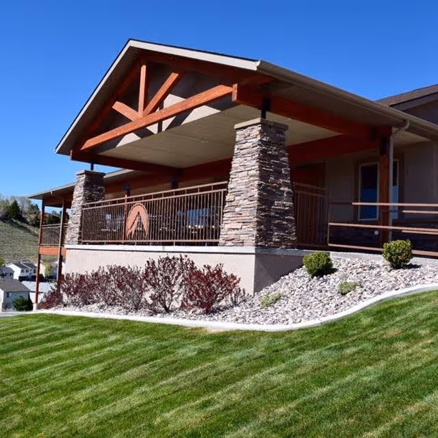 Exterior view of a building with a covered porch supported by stone pillars, surrounded by a well-maintained lawn and landscaping with bushes and rocks under a clear blue sky.
