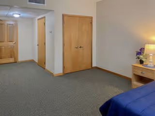 Interior view of a room with beige walls and carpeted floor. There are three wooden doors visible, one on the left wall, one in the center, and one slightly open on the far left. A small wooden nightstand with a lamp and a small flower arrangement is on the right side next to a blue bedspread.