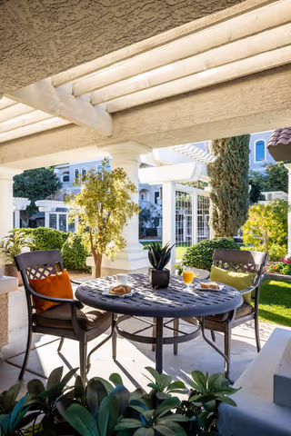 Outdoor patio area with a round metal table and two chairs, each with colorful cushions. On the table are a small plant, a glass of orange juice, and some pastries. The patio is shaded by a pergola, and there are green bushes, trees, and a white building in the background under a sunny sky.