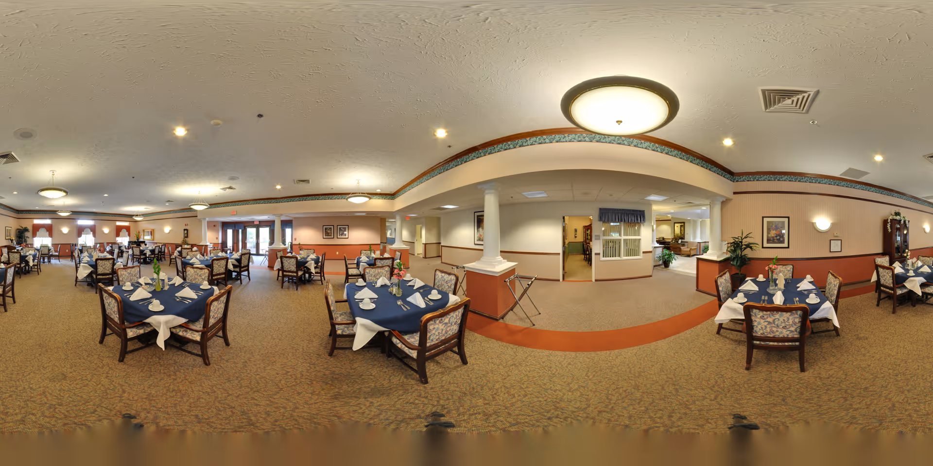 Spacious dining room with multiple round tables set with blue tablecloths and chairs in a senior living facility.