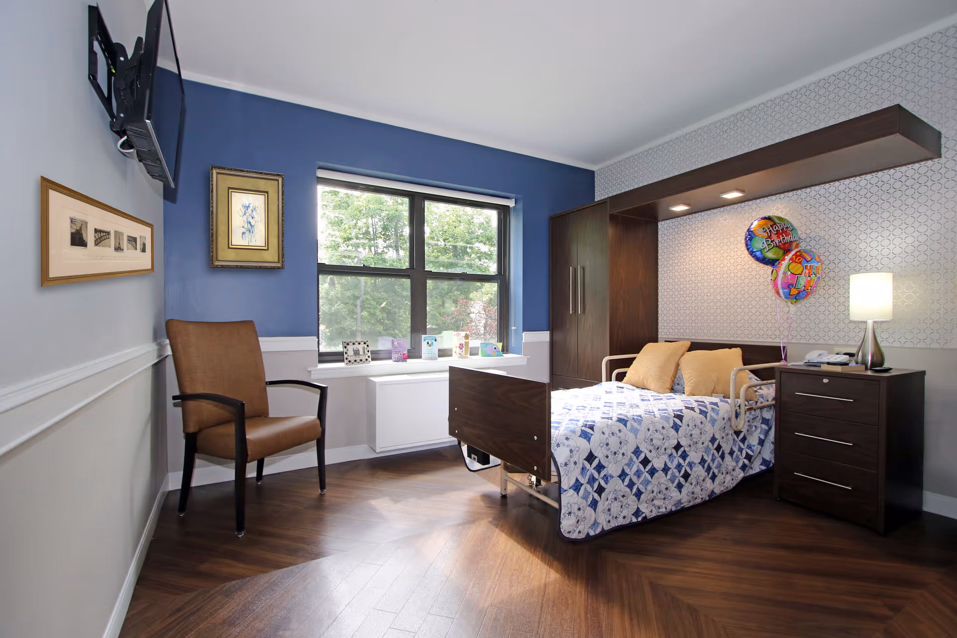 A bright bedroom in a senior living facility with a bed, nightstand, chair, wall-mounted TV and a window looking outside.