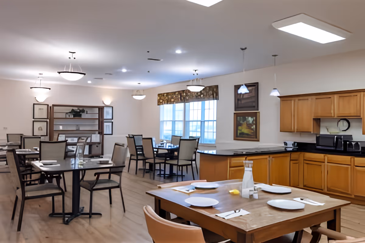 Bright communal dining room with multiple set tables and chairs and a kitchenette/service counter in the background.