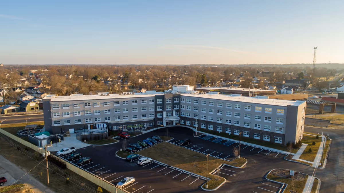 Aerial view of a four-story senior living building with a surrounding parking lot and nearby residential neighborhood.
