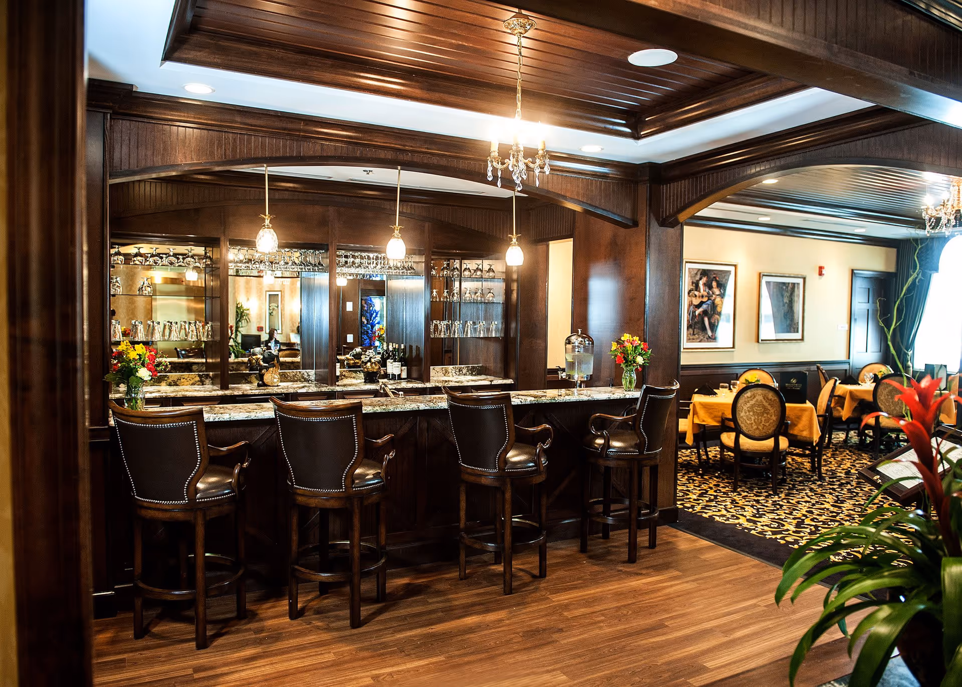Interior view of a senior living facility dining area featuring a wooden bar with four leather bar stools, pendant lights hanging above, and a mirrored back wall with glassware. Adjacent to the bar is a dining area with tables covered in yellow tablecloths and upholstered chairs. The room has wooden flooring, decorative ceiling beams, and framed artwork on the walls.