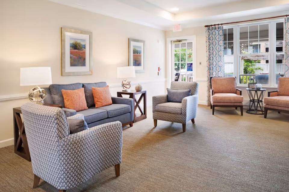 A bright and cozy living room area with a gray sofa adorned with orange cushions, two patterned armchairs with blue cushions, and two additional orange armchairs near a window with patterned curtains. The room features beige carpeting, two side tables with lamps, framed landscape artwork on the walls, and a glass door leading outside.