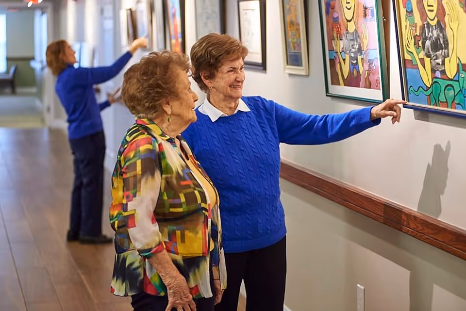 Two elderly women standing in a hallway looking at colorful framed artwork on the wall. One woman is pointing at a piece of art while the other watches. In the background, another person is adjusting or hanging artwork further down the hallway.