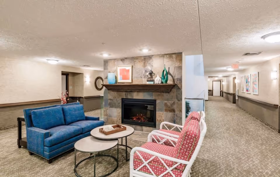 A cozy seating area in a hallway of a retirement community featuring a blue couch, two white chairs with red patterned cushions, a round coffee table, and a fireplace with decorative items on the mantel. The hallway is carpeted and has framed artwork on the walls.