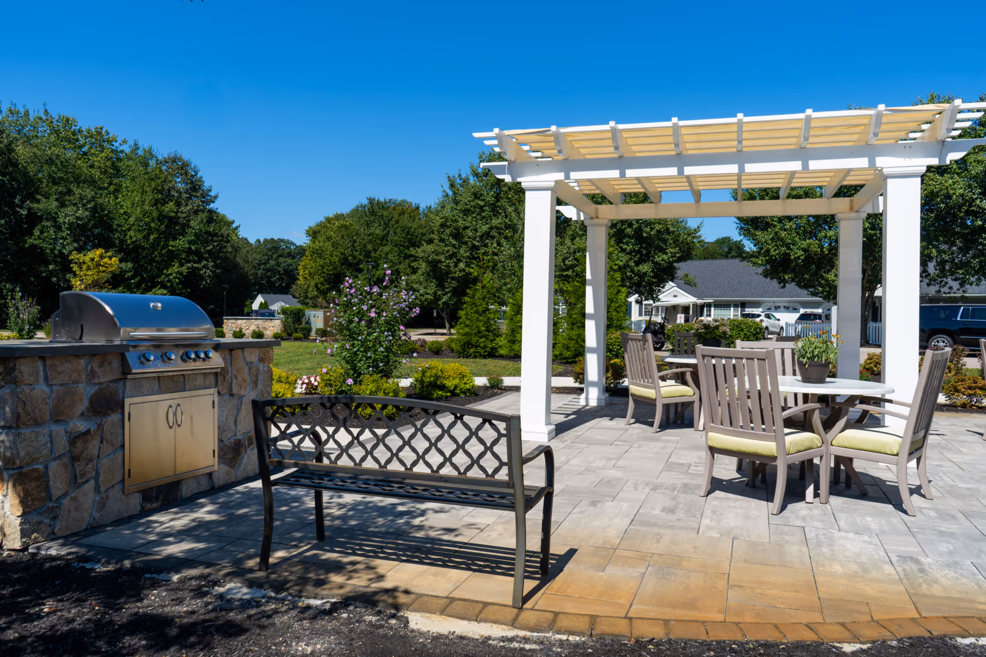 Outdoor patio with a white pergola, dining table and chairs, a bench, and a built-in grill set among landscaped grounds.