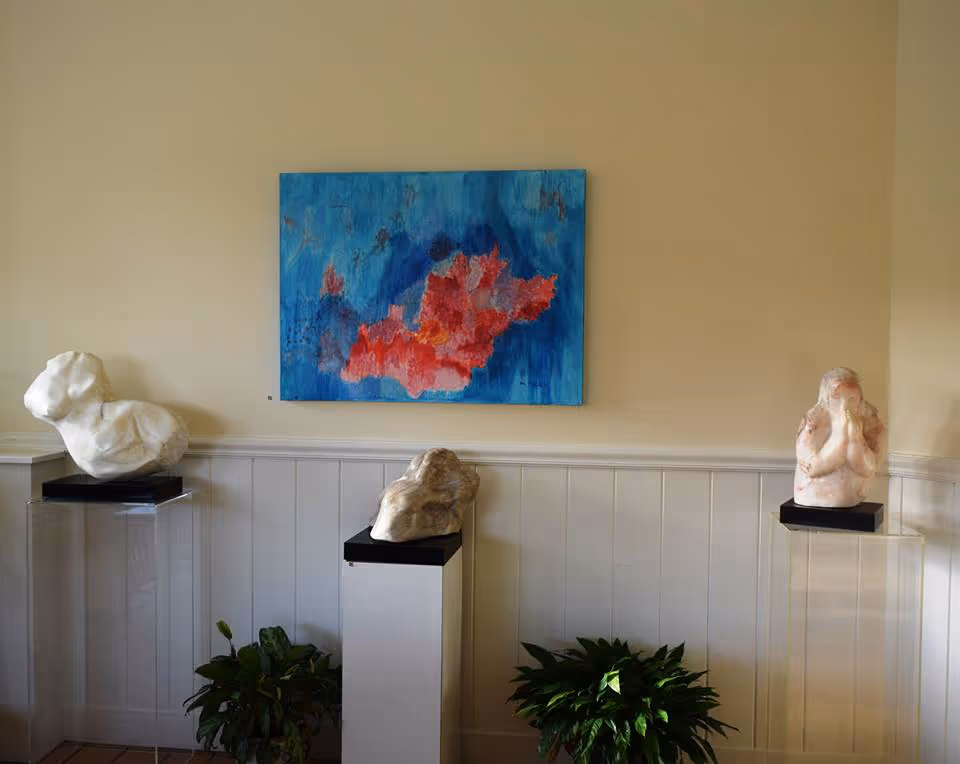 An interior room with three sculptures displayed on pedestals against a beige wall with white wainscoting. Above the sculptures hangs a colorful abstract painting with blue and red tones. Two green plants are placed on the floor beneath the pedestals.
