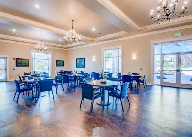 Bright community dining room with round tables and blue chairs under chandeliers and large windows.