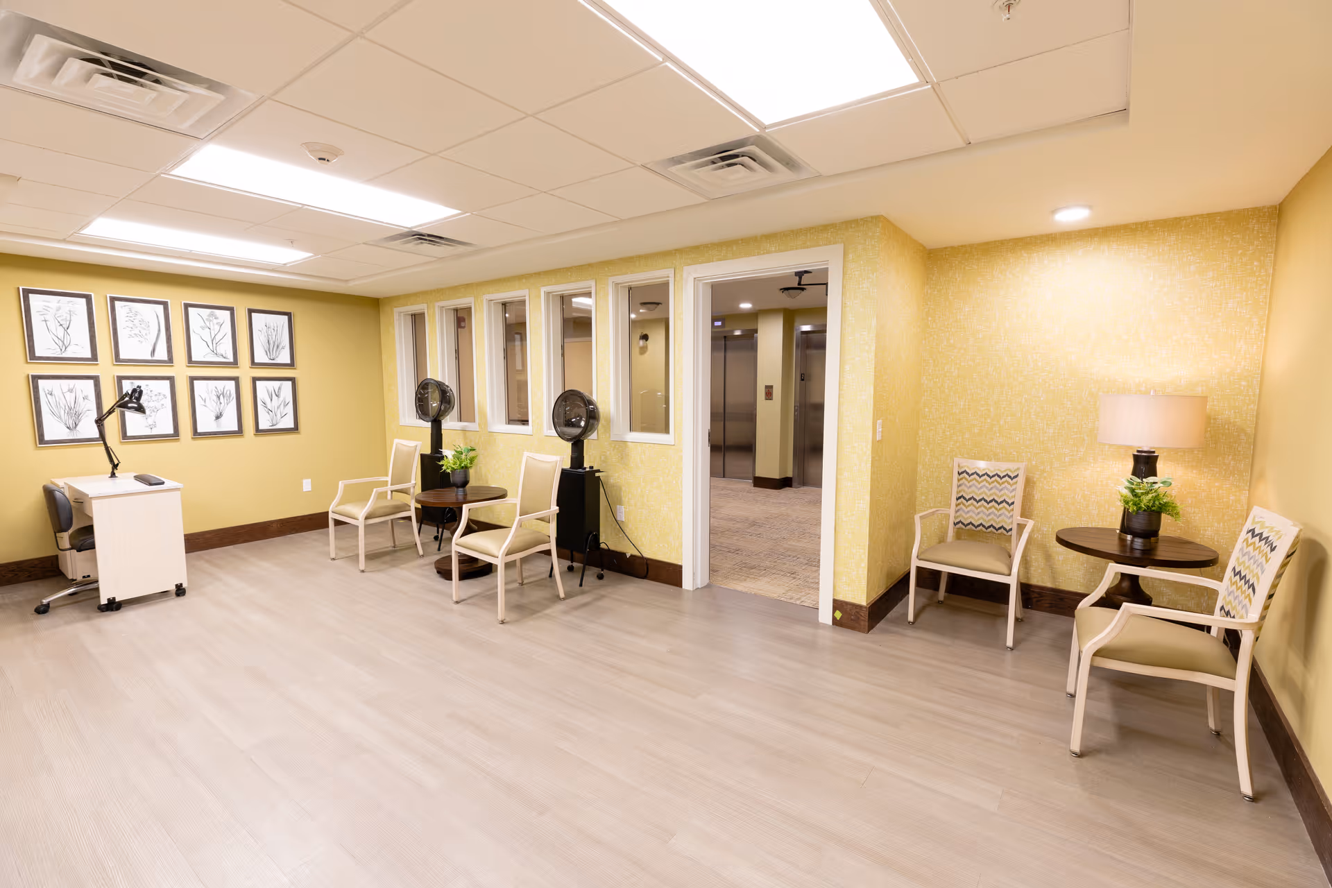 A well-lit senior living facility common area with light wood flooring and yellow walls. The room features several chairs arranged around small tables with potted plants and a lamp. On one wall, there are framed botanical prints, and a small desk with a lamp and chair is positioned nearby. A doorway leads to a hallway with elevators visible in the background.