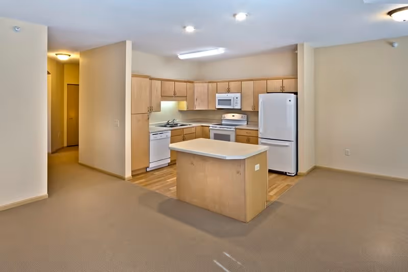 A clean, modern kitchen area in a senior living facility with light wood cabinets, a white refrigerator, stove, microwave, and dishwasher. There is a small kitchen island in the center and beige carpet flooring surrounding the kitchen area with wood flooring in the kitchen space. The walls are painted a light beige color.