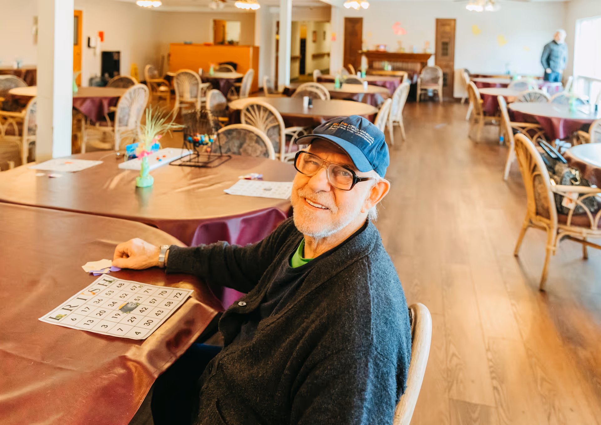An elderly man wearing glasses and a cap sits at a table in a large dining room with multiple tables and chairs. He is smiling and appears to be playing bingo, with a bingo card and markers on the table in front of him. The room has wooden flooring and is well-lit with natural light coming from windows on the right side.