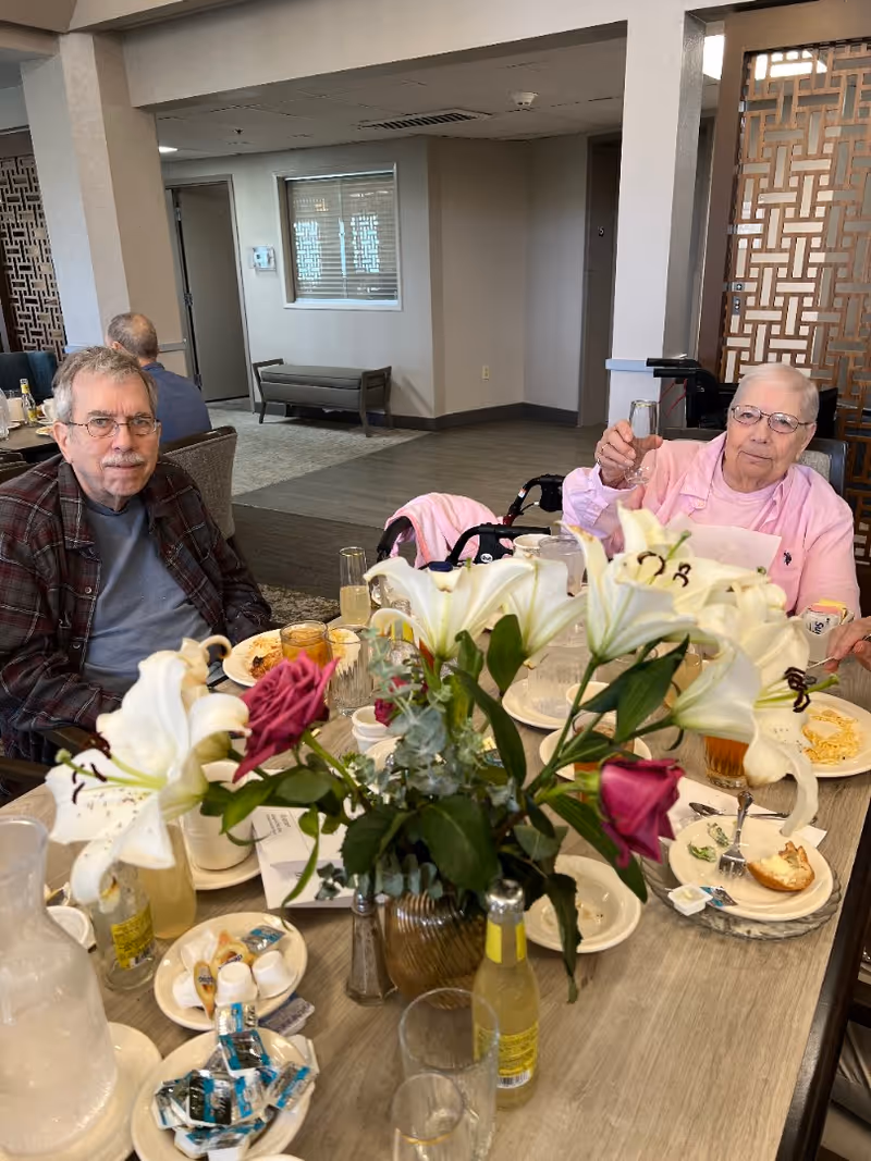 Two older adults seated at a dining table with plates, drinks, and a large floral centerpiece in a communal dining area.