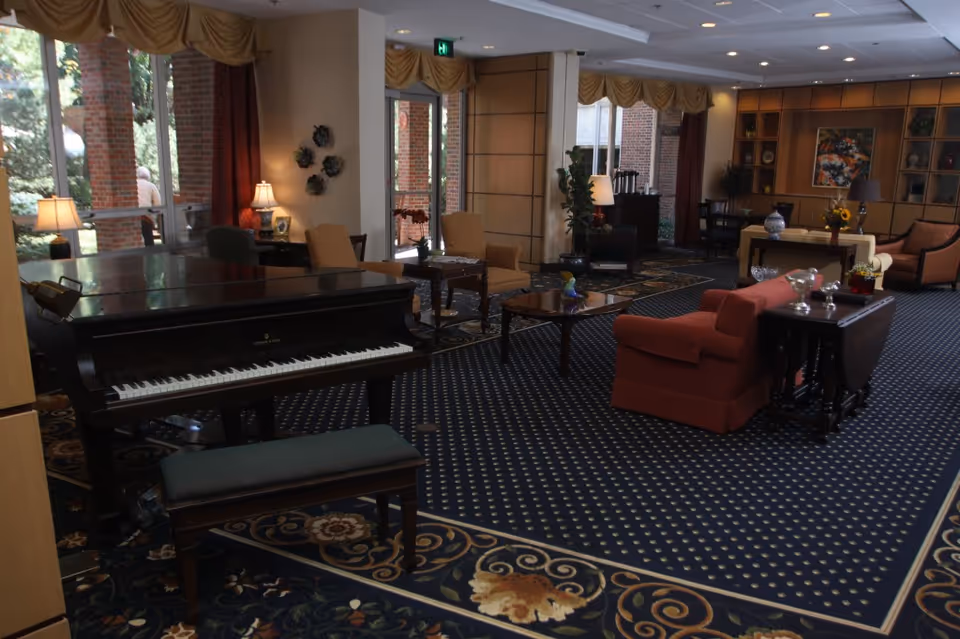 A spacious and elegantly furnished common area in a retirement community featuring a grand piano with a bench, multiple seating arrangements including armchairs and sofas, decorative lamps, a patterned carpet, and large windows with curtains allowing natural light to enter. There are also plants and artwork on the walls, creating a warm and inviting atmosphere.
