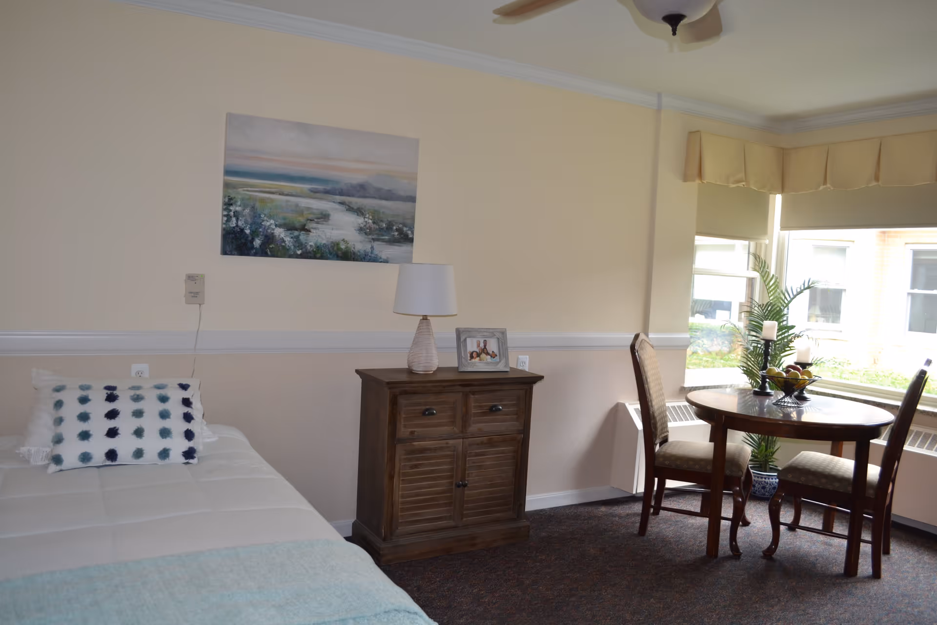 A cozy bedroom in a senior living facility with a single bed on the left side, a wooden nightstand with a lamp and a framed photo on top, a landscape painting on the wall, and a round wooden table with two chairs near a large window with beige valances. A potted plant and decorative candles are on the table, and natural light comes through the window.
