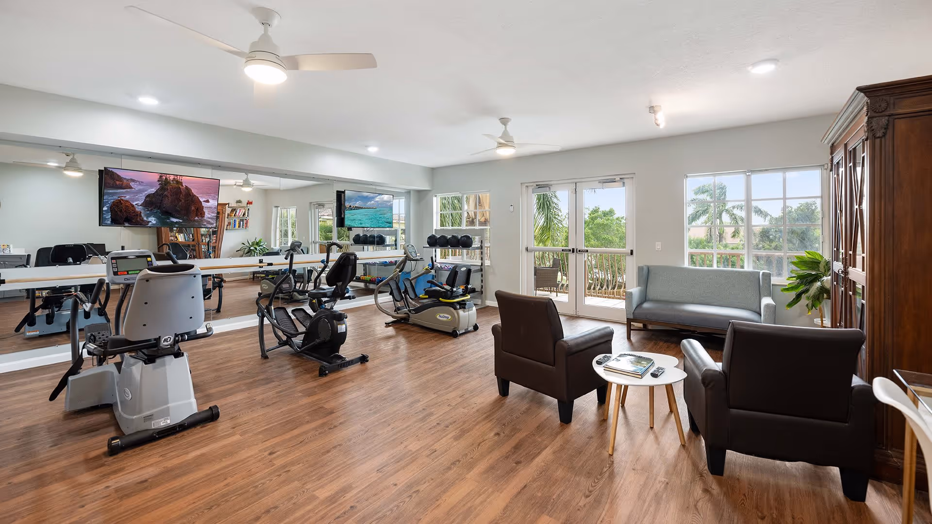 A bright exercise room with wooden flooring featuring several stationary exercise bikes and a large wall mirror. There is a seating area with two brown armchairs, a small round table, and a light blue sofa near large windows and glass doors that open to a balcony with a view of palm trees outside. A large wooden cabinet and some plants are also visible in the room.
