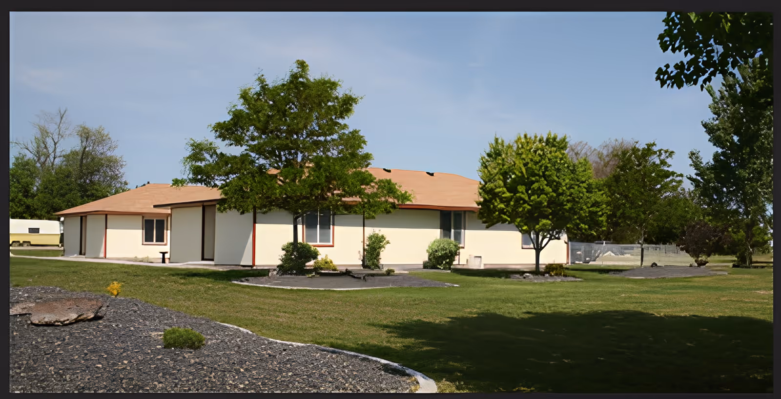 Single-story building with a light beige exterior and a reddish-brown roof surrounded by green grass, trees, and landscaped areas under a clear blue sky.