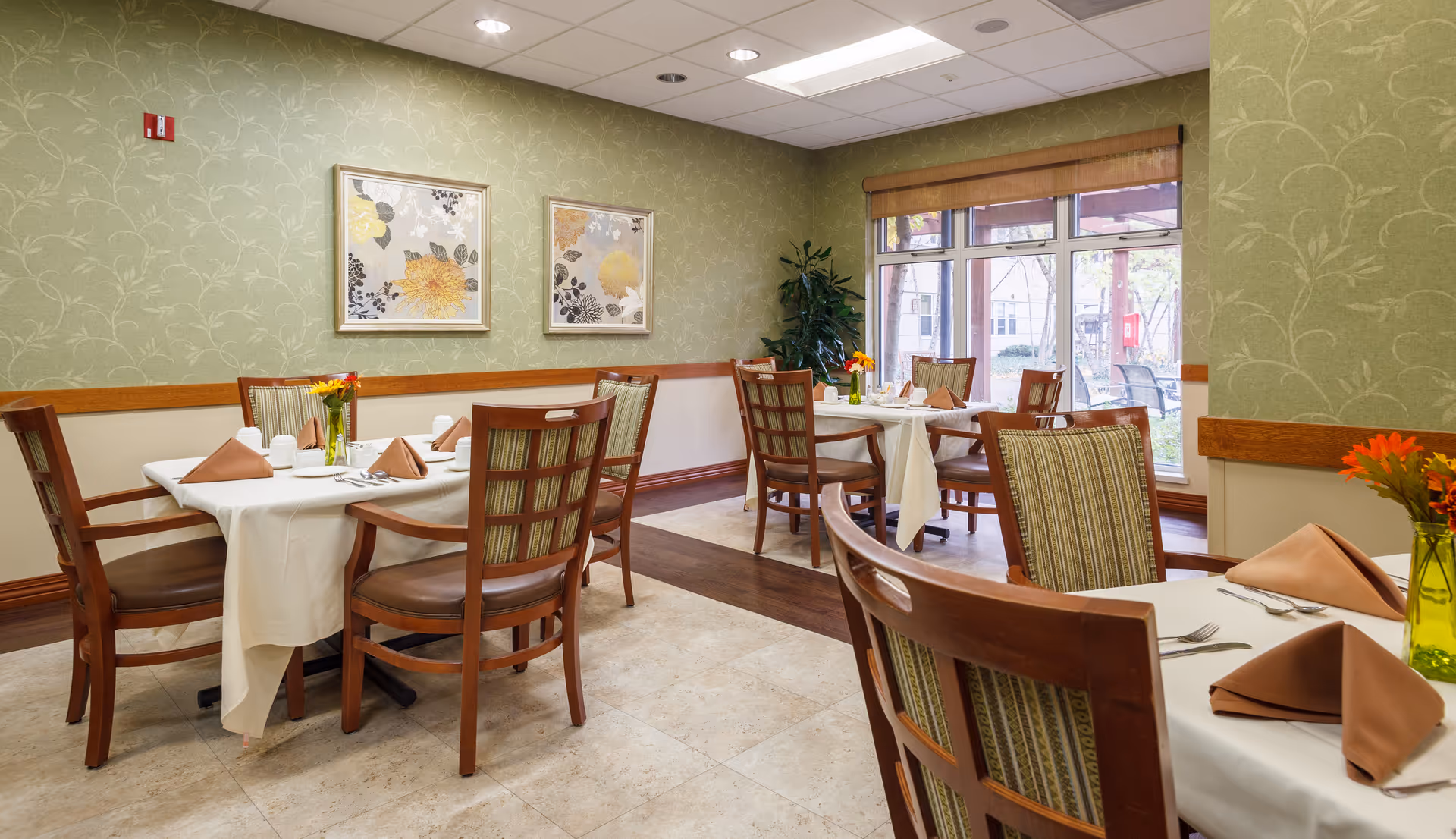 A dining room with several tables covered in white tablecloths, each set with brown folded napkins, silverware, and small flower vases. The room has green patterned wallpaper, two framed floral paintings on the wall, wooden chairs with striped cushions, and large windows letting in natural light.