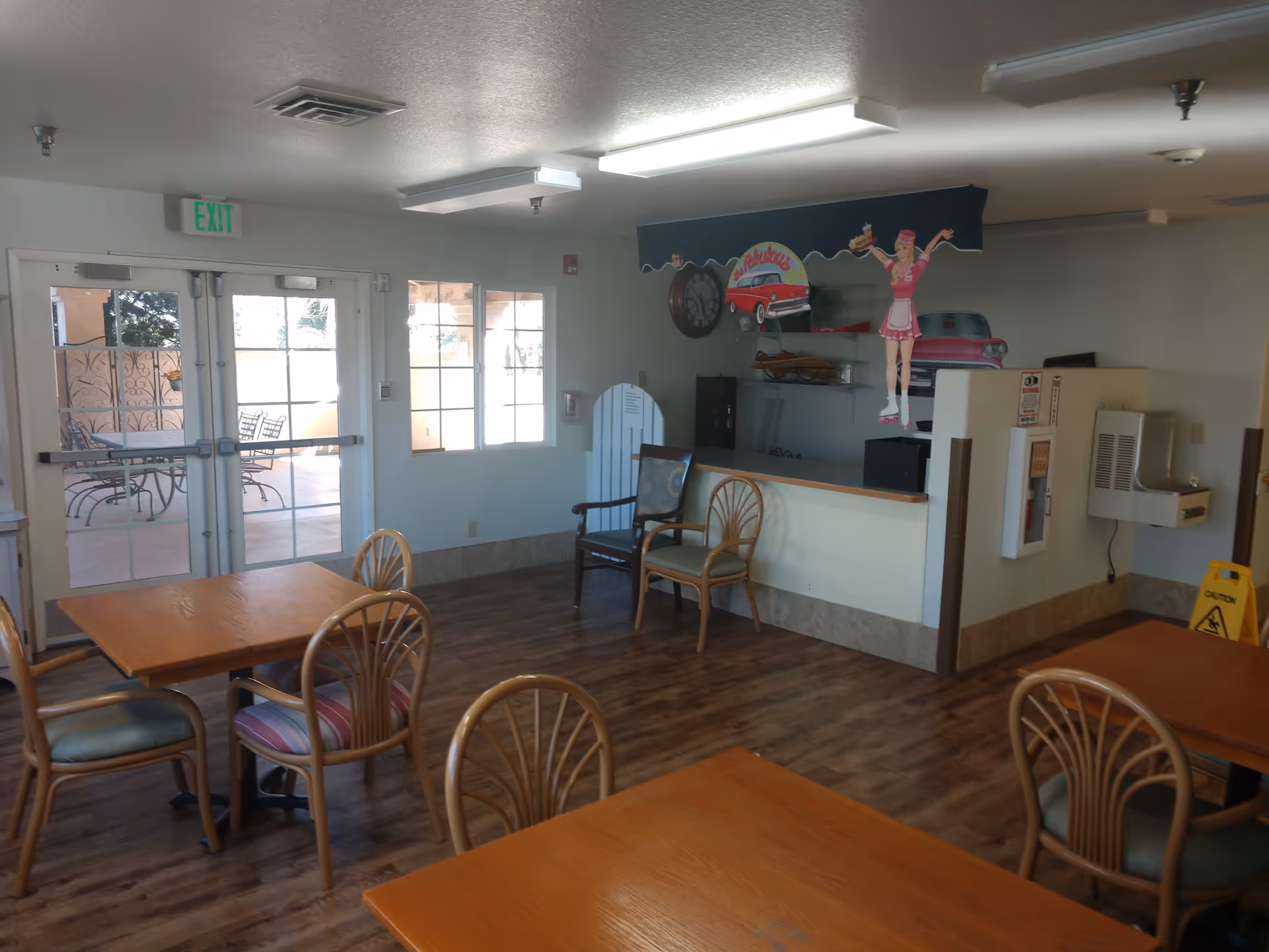 Interior view of a dining area with wooden tables and chairs, a water fountain, and a counter decorated with retro diner-themed wall art including a waitress on roller skates and vintage cars. There are double glass doors leading to an outdoor patio with metal chairs and tables.