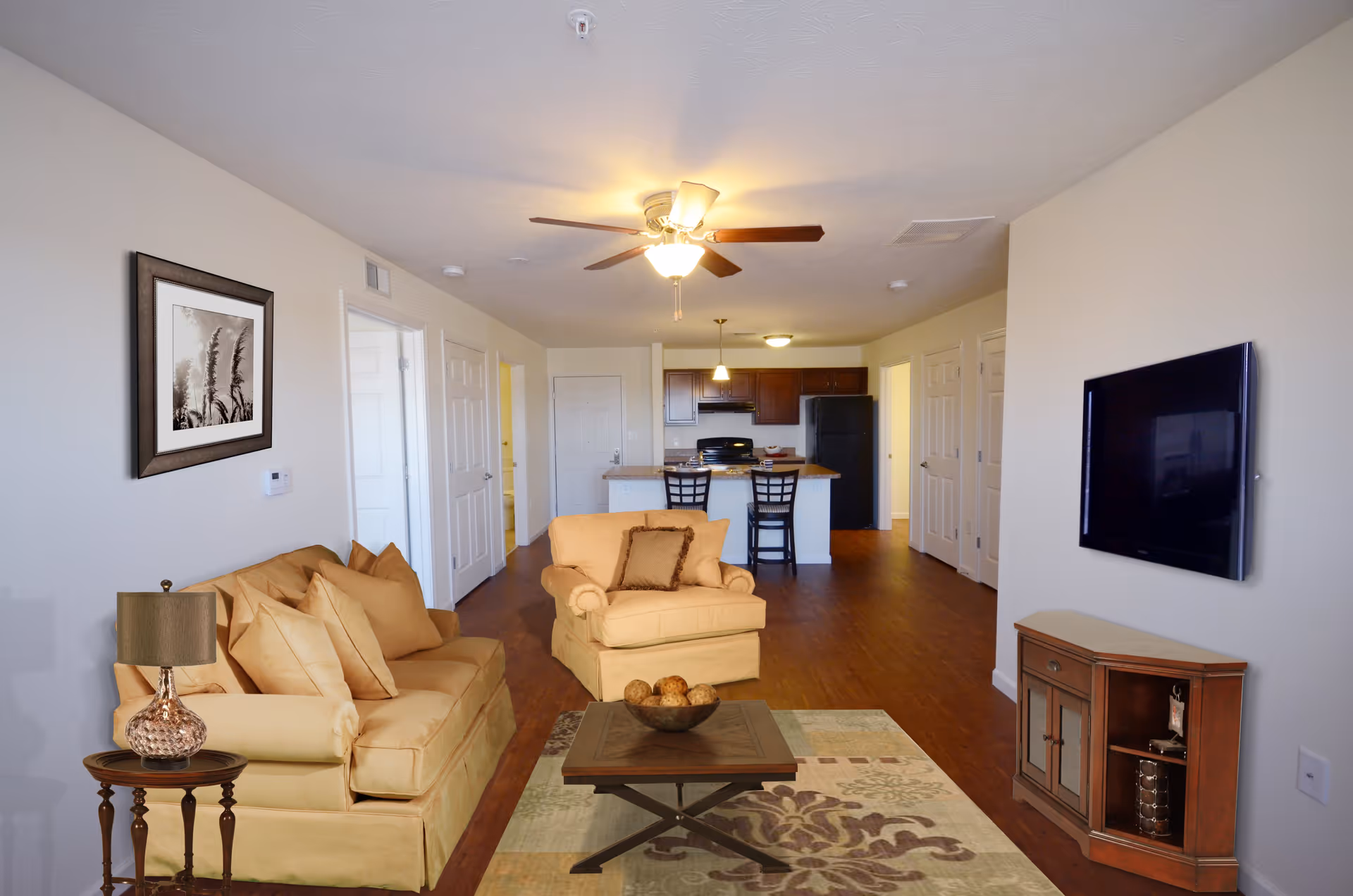 A living room area with beige sofas and a matching armchair, a wooden coffee table with decorative balls, a side table with a lamp, a wall-mounted flat-screen TV, and a framed picture on the wall. The room has wooden flooring and a ceiling fan with a light. In the background, there is an open kitchen with dark wood cabinets, a black refrigerator, and a kitchen island with two chairs.