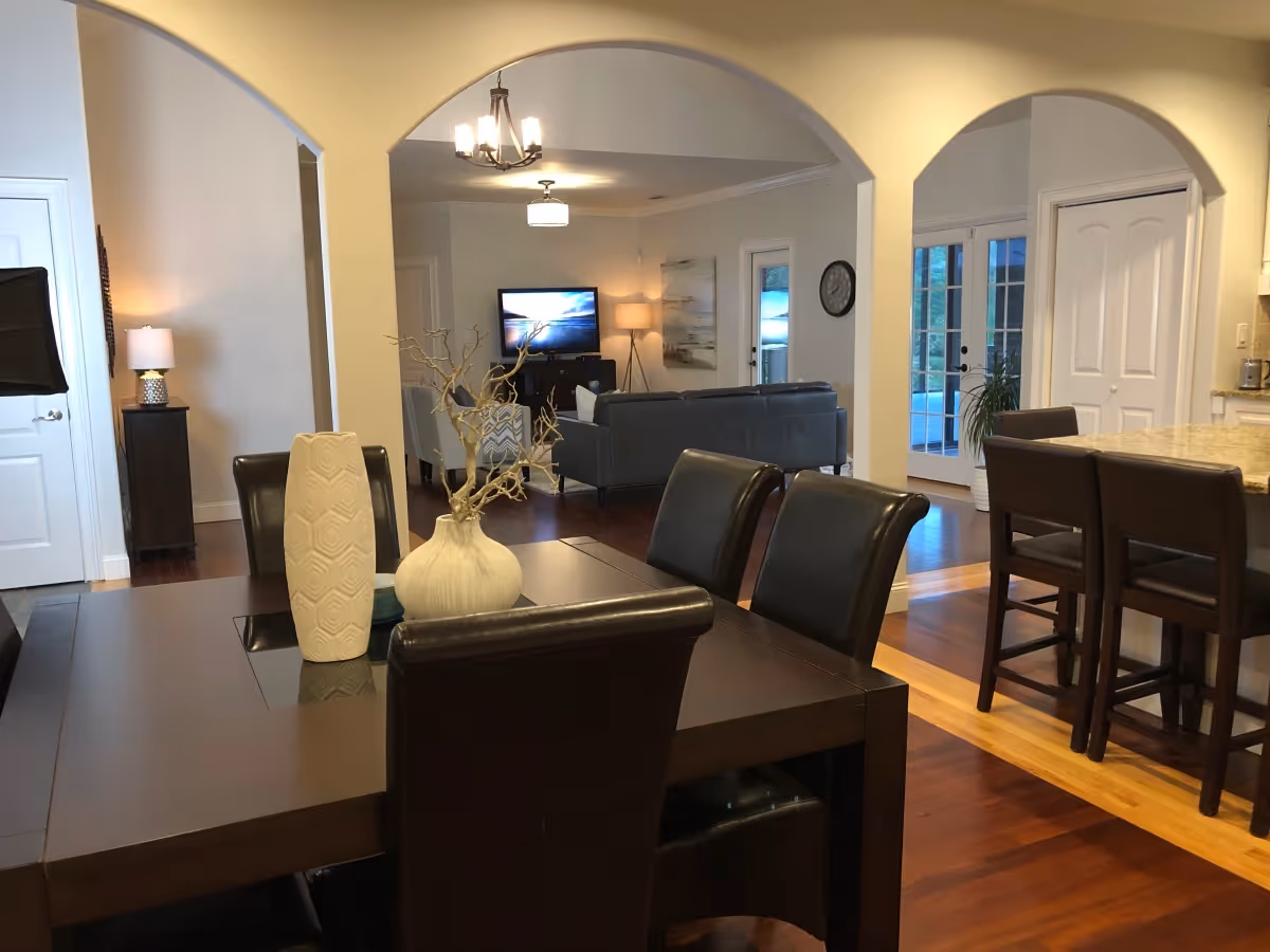 Interior view of a senior living facility showing a dining area with a dark wooden table and leather chairs, decorated with vases and a branch centerpiece. In the background, there is a living room with a TV, sofas, and a floor lamp. The space features arched doorways, hardwood floors, and a kitchen area with bar stools on the right side.