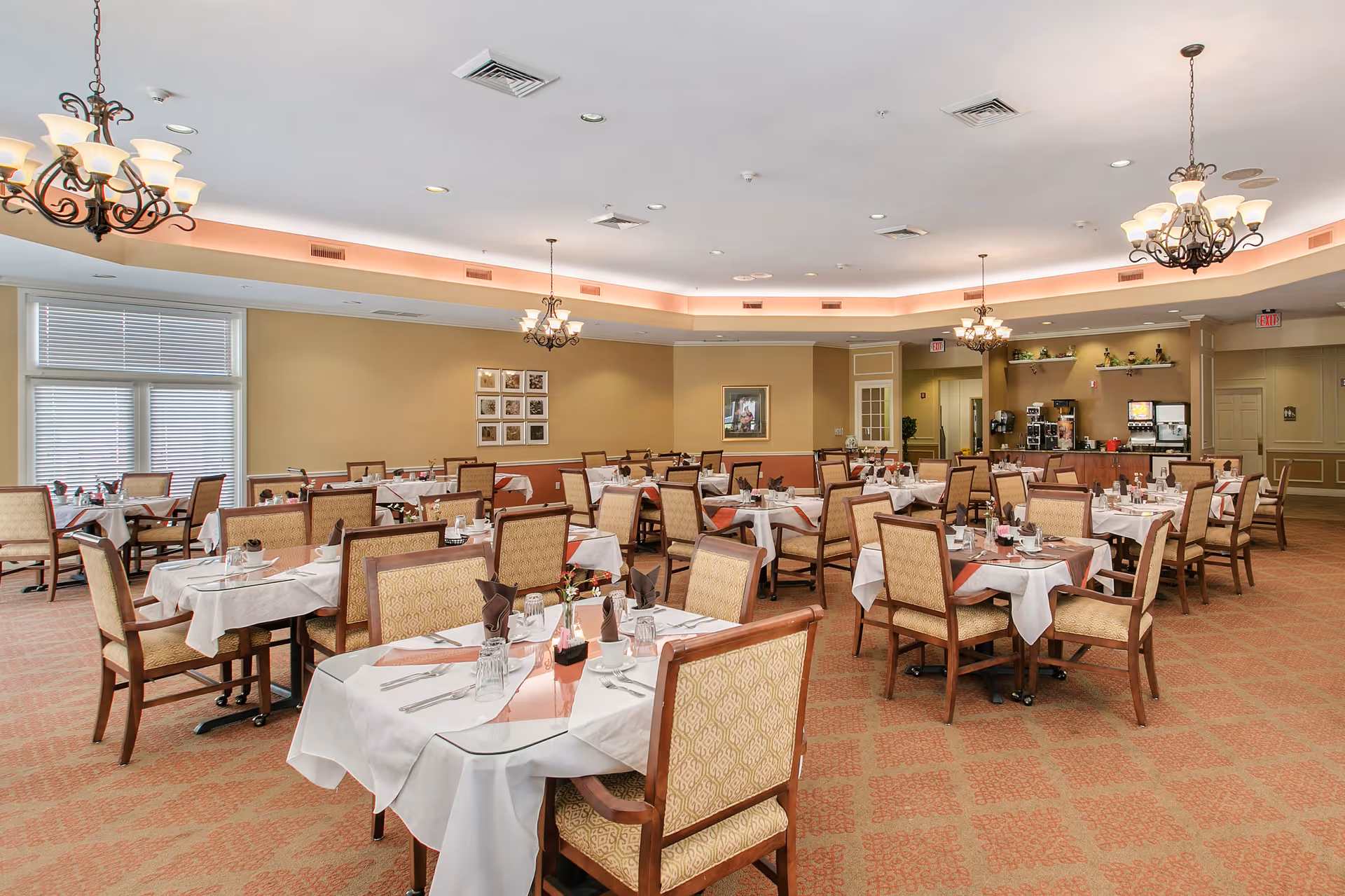 A spacious dining room with multiple tables covered in white tablecloths and set with glasses, napkins, and silverware. The room features beige walls, patterned carpet, large windows with blinds, and several ornate chandeliers hanging from the ceiling. There is a beverage station with coffee machines and dispensers in the background.