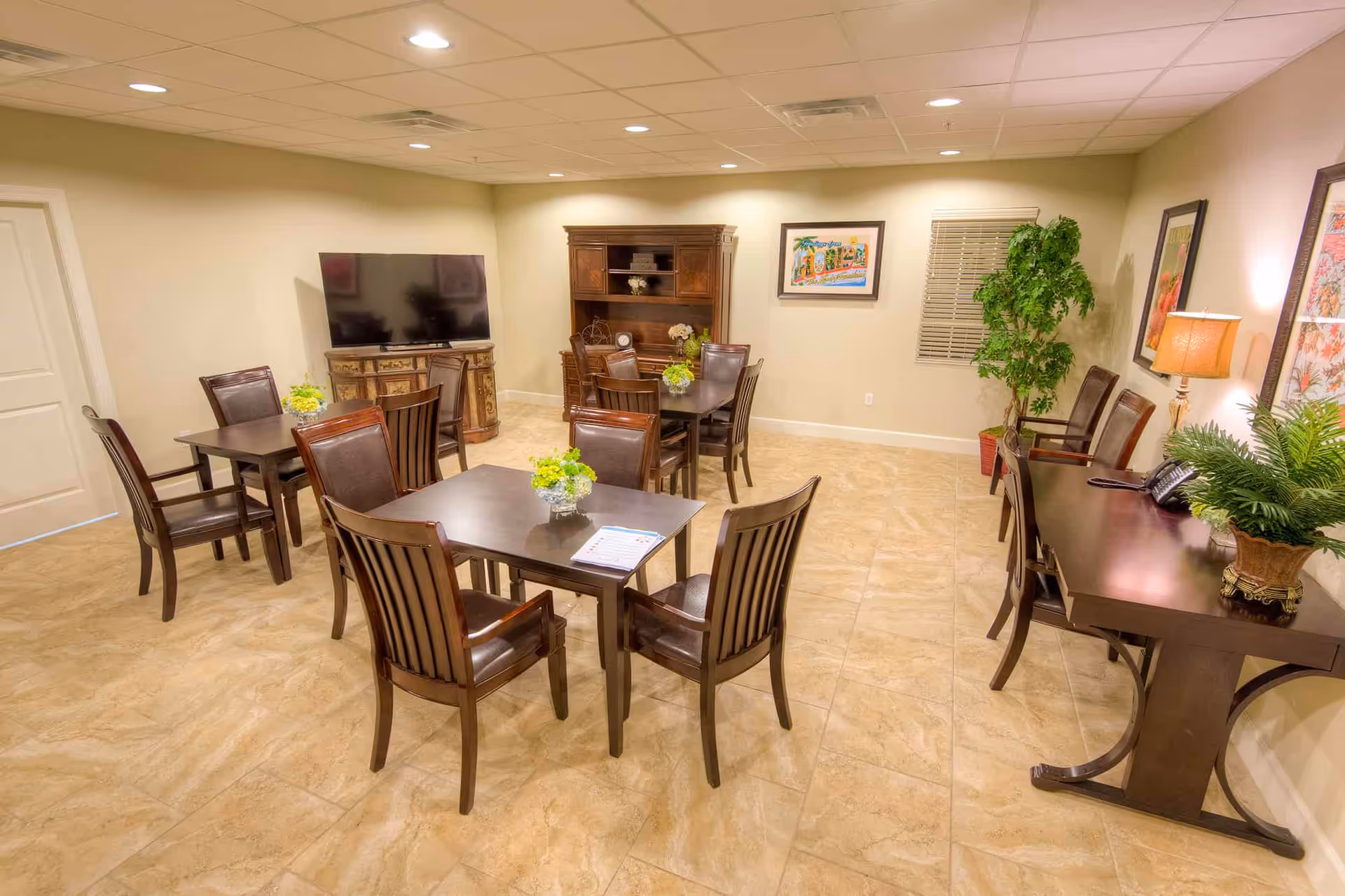 A well-lit common room with several dark wooden tables and chairs arranged neatly. There is a large flat-screen TV on a decorative cabinet against the wall. The room also features a wooden hutch, framed artwork, a potted plant, and a side table with a lamp and greenery. The floor is tiled with a beige marble pattern.