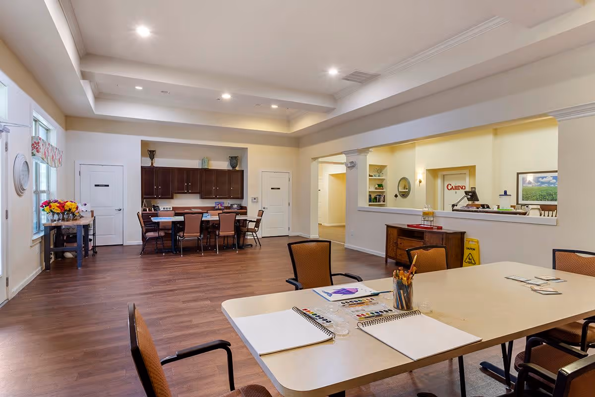 A spacious, well-lit common area in an assisted living facility with a large table in the foreground set up for arts and crafts, including paint palettes, brushes, and paper. In the background, there is a dining table with chairs, cabinets, and a side table with flowers near a window. The room has wood flooring, recessed lighting, and light-colored walls.