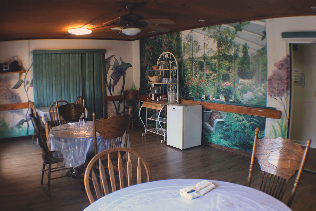 Communal dining room with round tables and wooden chairs, a small refrigerator and a large garden mural on the wall.