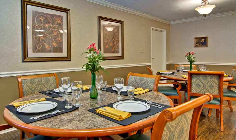 Well-set dining room with round tables, place settings, floral centerpieces, and framed artwork on the walls.